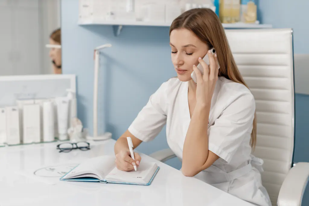 A woman in a white tunic at a white desk, a phone held to her ear, writing in an open diary with skincare products on the shelves behind her.