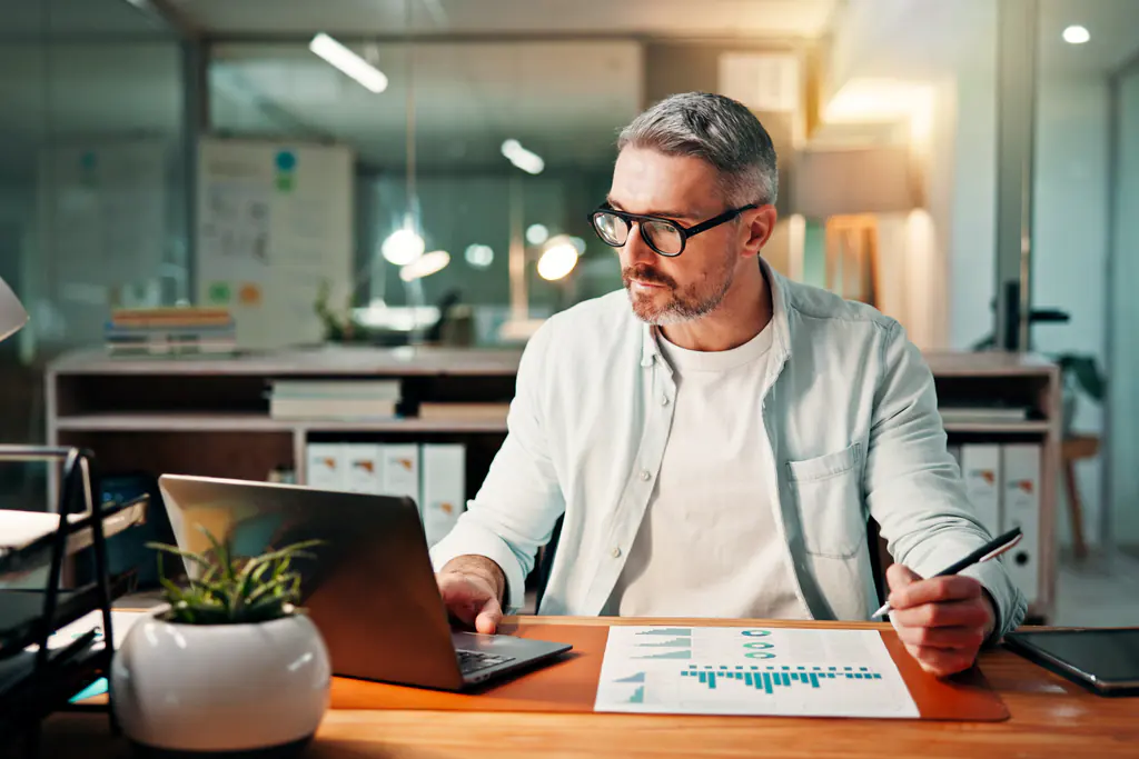 A man in glasses at a desk in the evening, a printed sheet of bar charts in front of him and a laptop open to one side.