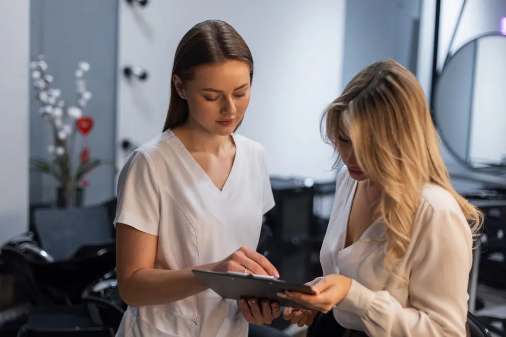 Two women looking down at a clipboard held between them in a softly lit salon interior. The woman on the left wears a white tunic.