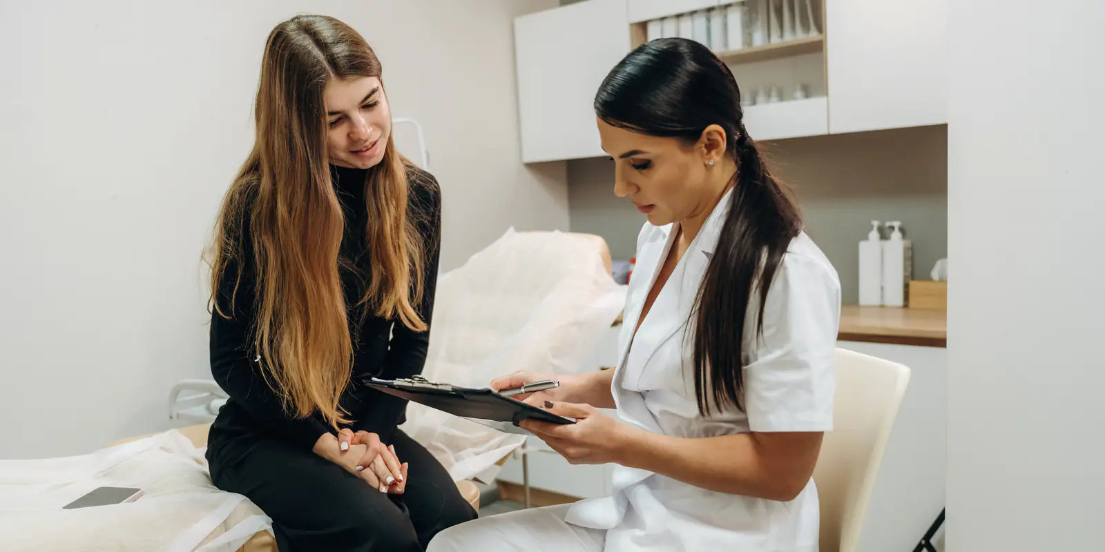A practitioner in a white uniform seated beside a patient in a treatment room, going through a form on a clipboard together.