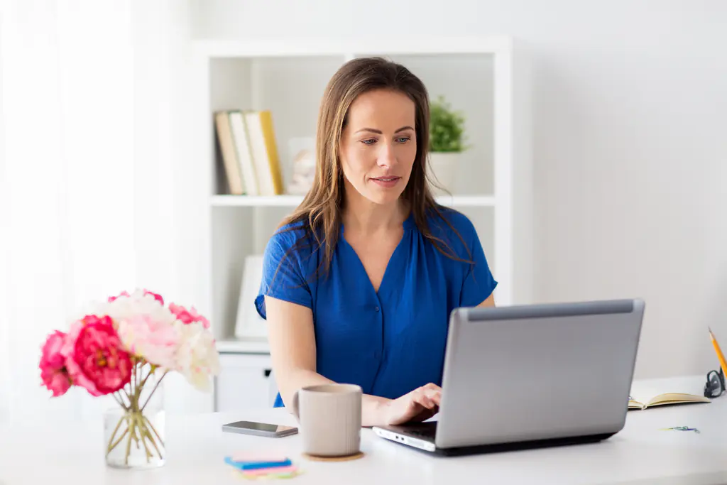 A woman in a blue top working at a laptop at a white desk, with a vase of pink flowers, a mug and a phone beside her.