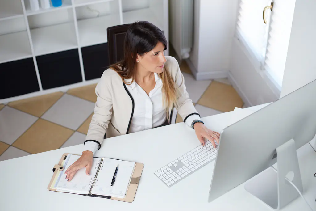 Seen from above, a woman at a white desk types at a keyboard with an open paper diary and a pen beside her.