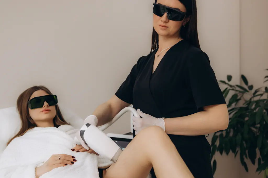 A practitioner in protective goggles performing a laser treatment on a patient’s leg in a clinic room.
