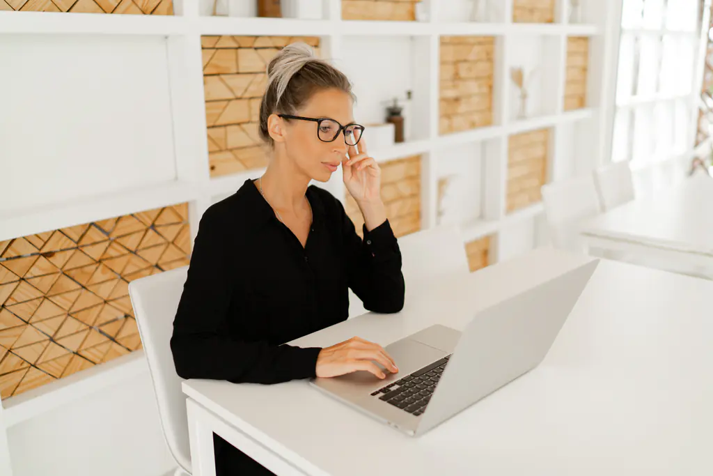 A woman in a black shirt and glasses working at a laptop on a white table, a wall of timber shelving behind her.