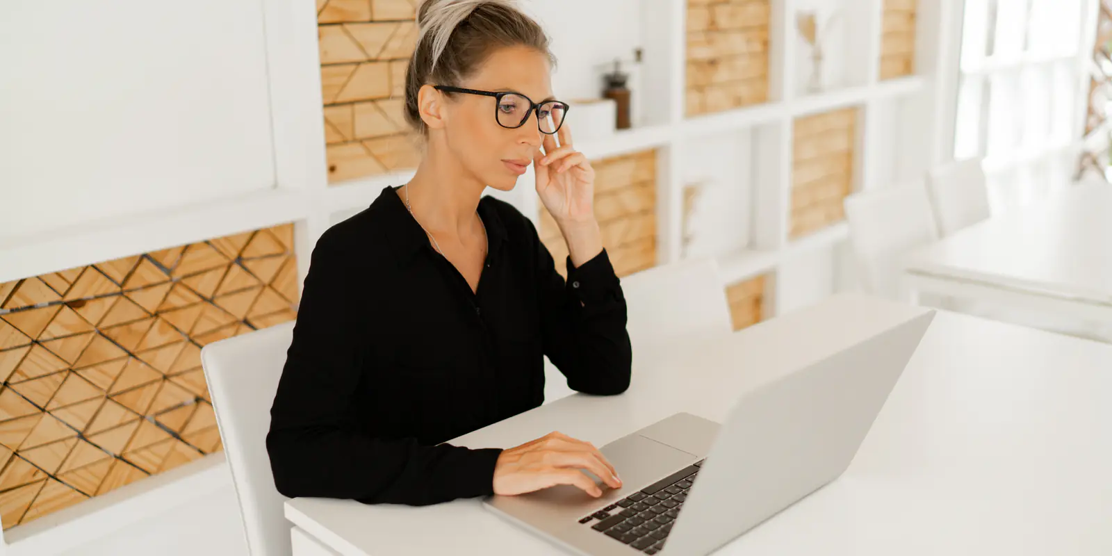 A woman in a black shirt and glasses working at a laptop on a white table, a wall of timber shelving behind her.
