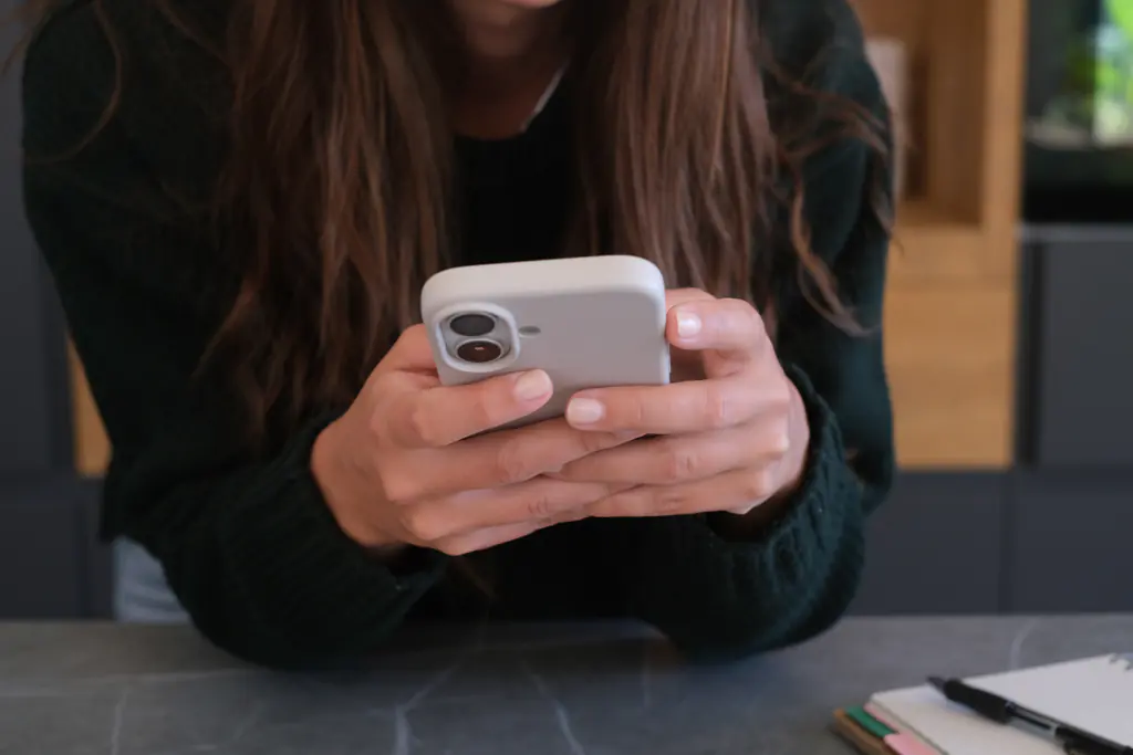 Close-up of a person’s hands typing on a smartphone at a stone counter in a warm interior.