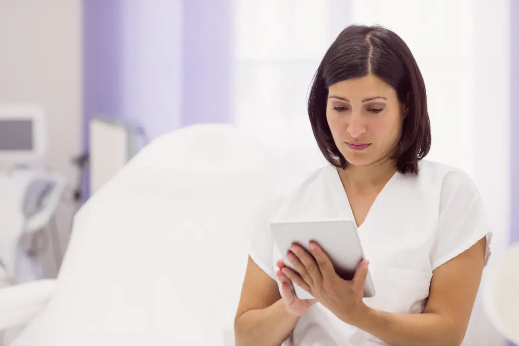 A woman in a white tunic reading a tablet held in both hands, a white treatment couch and soft lilac light behind her.