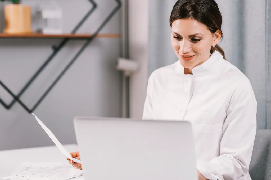 A woman in a white uniform at a laptop, reading a sheet of paper, with more paperwork on the desk in front of her.
